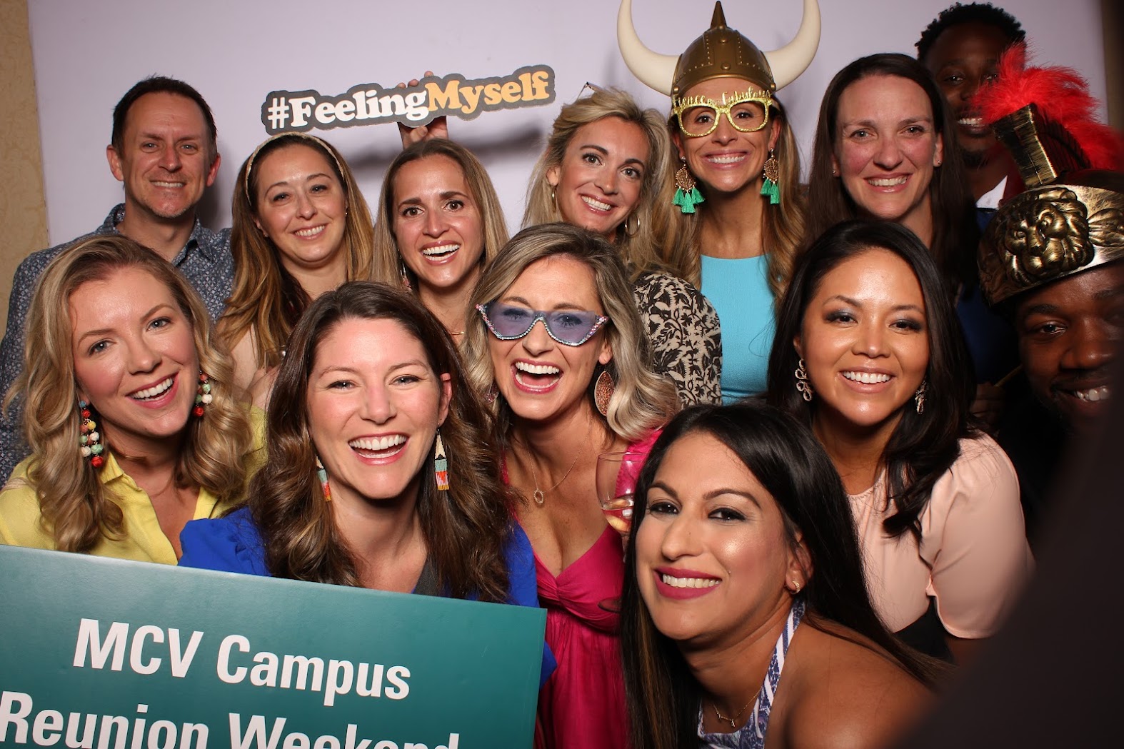 A group of people posing for a fun photo booth photo holding a sign that reads MCV Campus Reunion Weekend